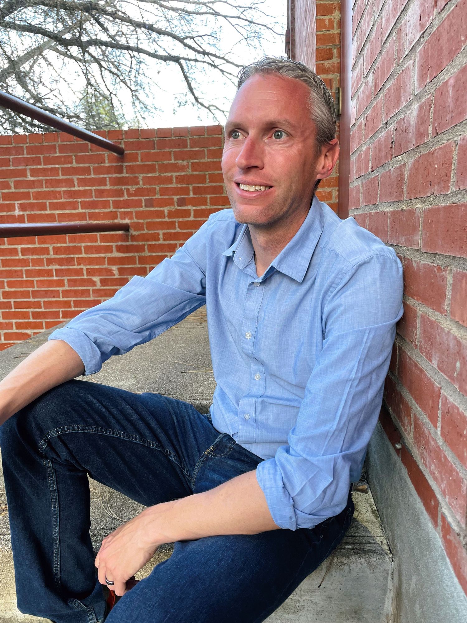 Casual portrait by brick wall wearing light blue button-down shirt and jeans.