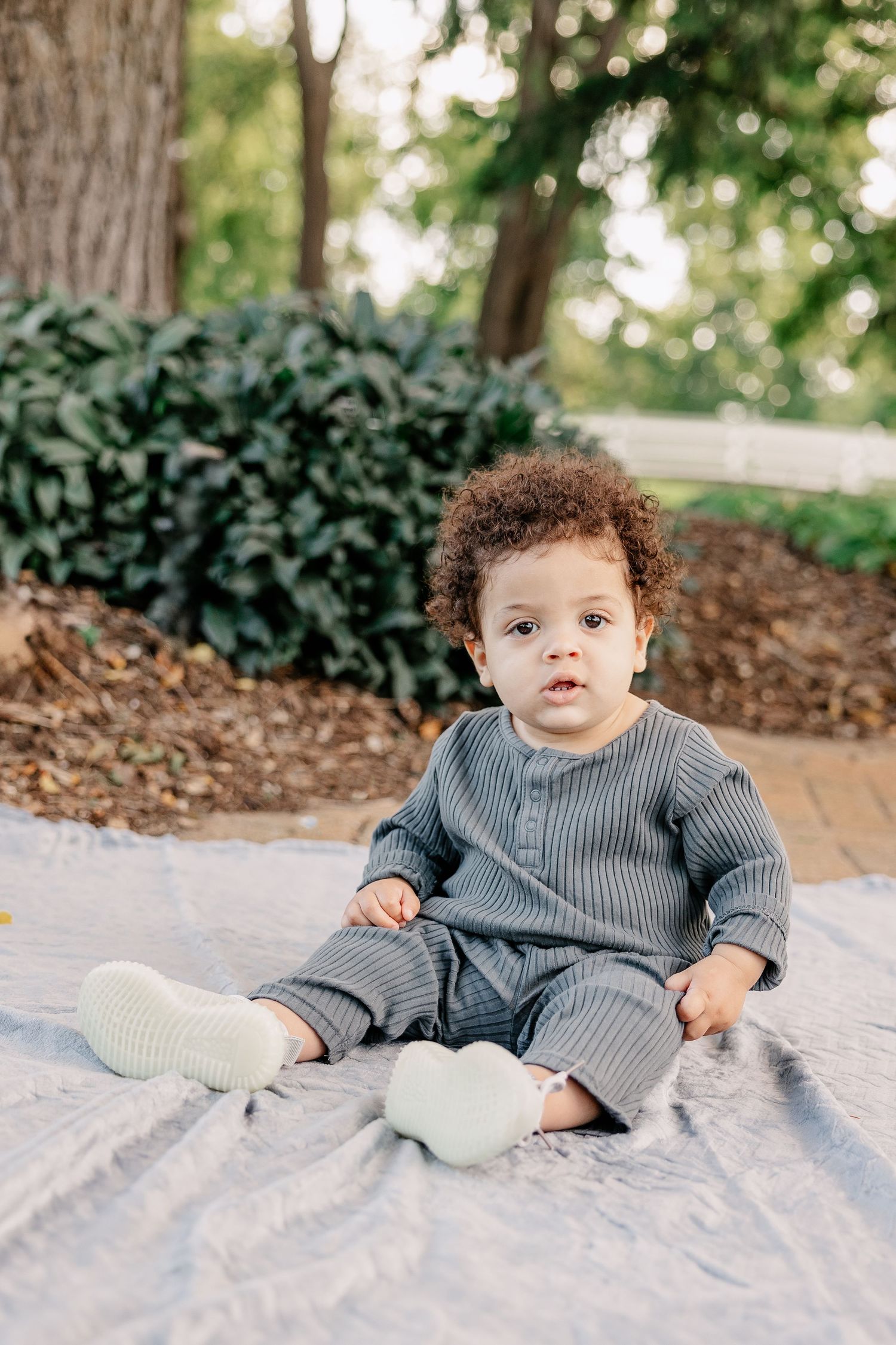 Small child in gray ribbed outfit sits on blanket in outdoor garden setting.