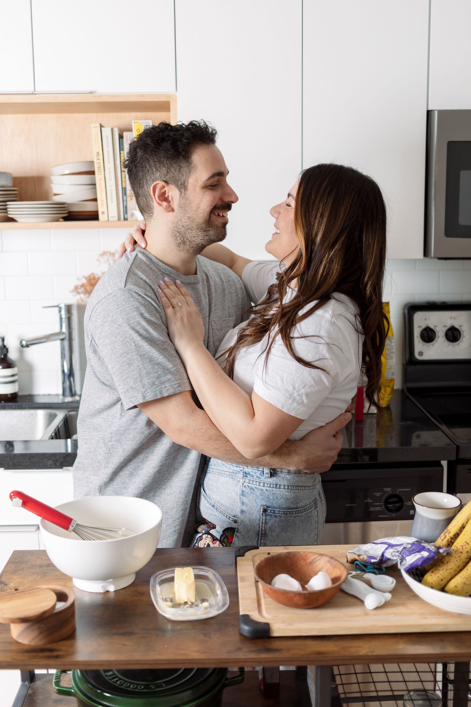 A couple embraces while cooking together in their kitchen with ingredients spread on the counter.