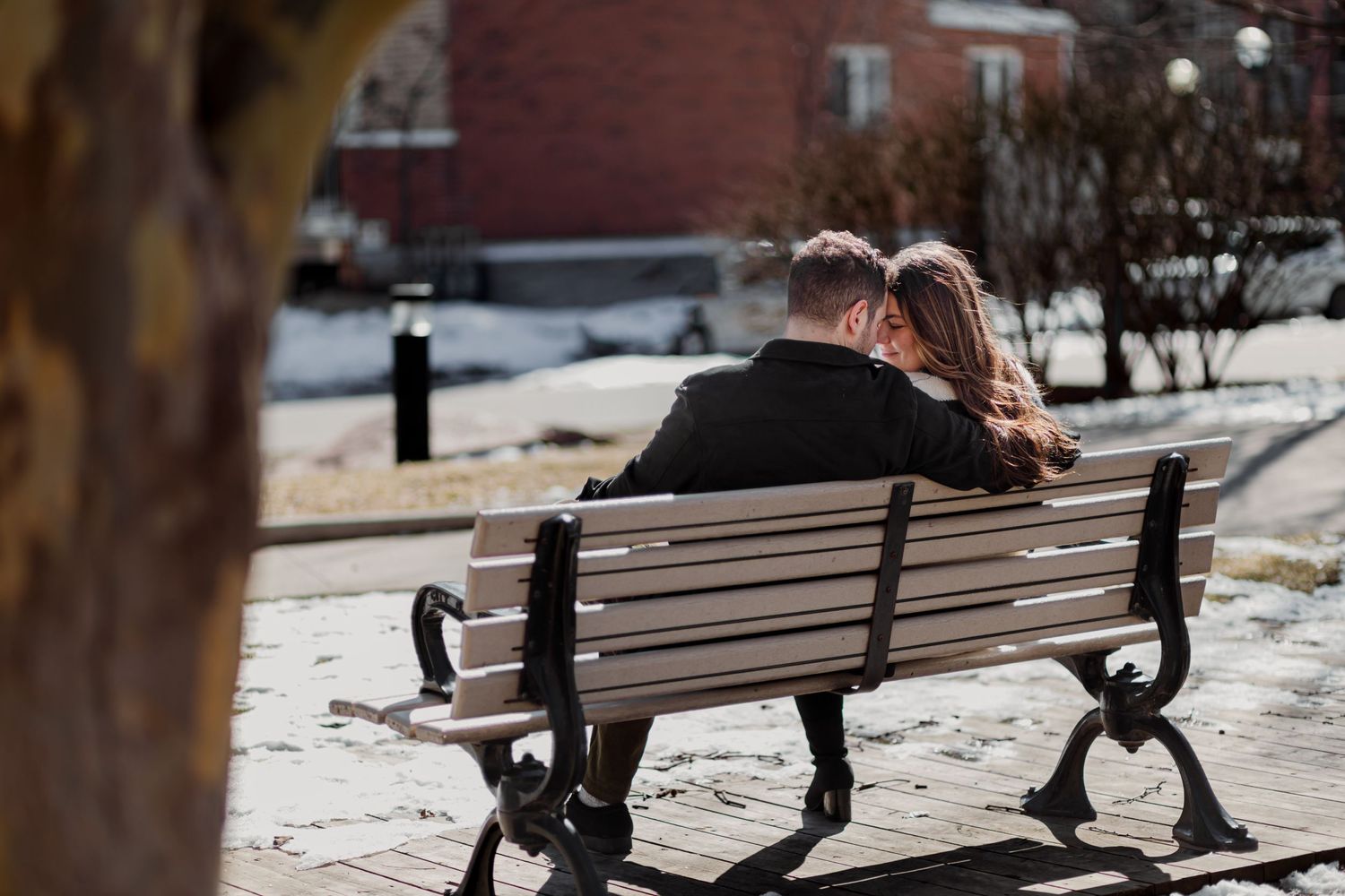 A couple sits close together on a wooden park bench on a winter day with snow on the ground.