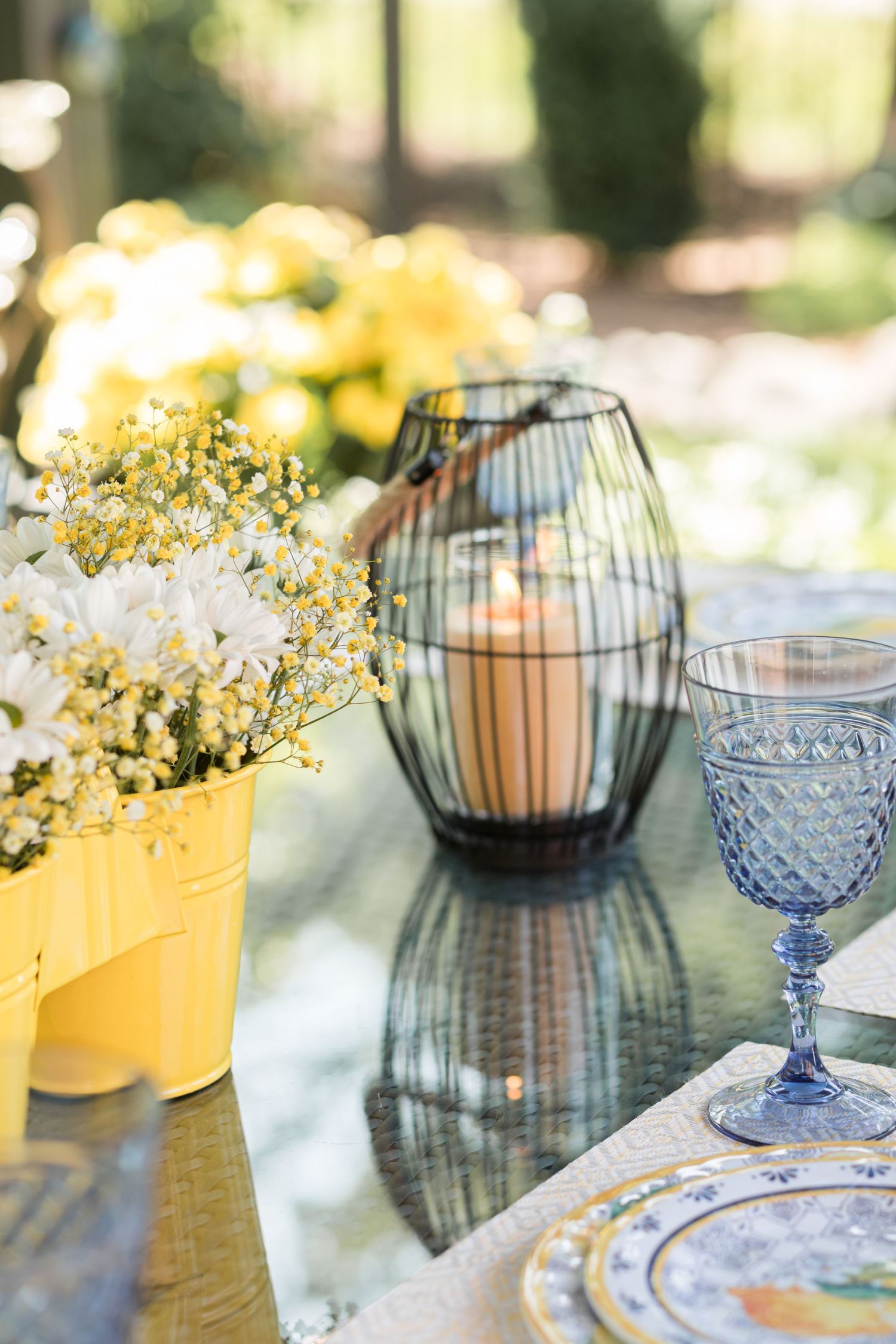 Table setting with blue vintage glasses, candle lanterns and yellow flowers in outdoor lighting.