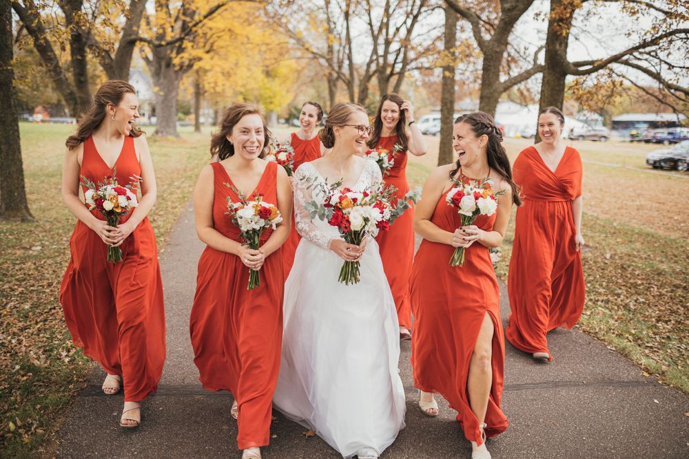 Bride and bridesmaids in flowing red dresses walking together on an autumn path.