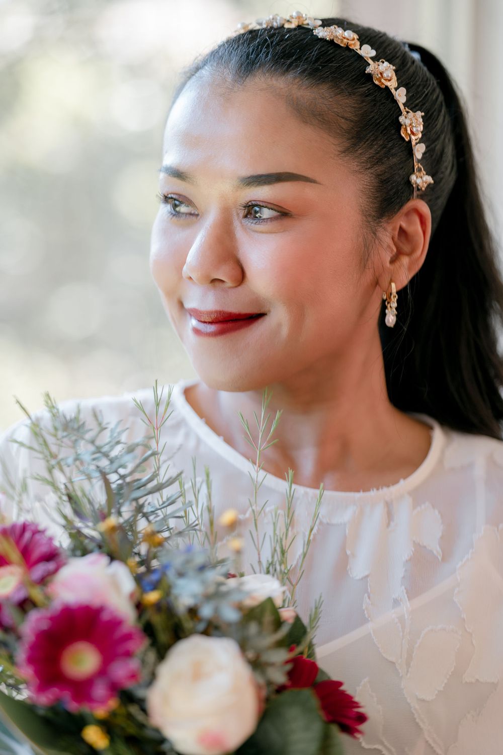 Mature asian bride in natural pink toned makeup  with jewelled headband holding bouquet and looking out window
