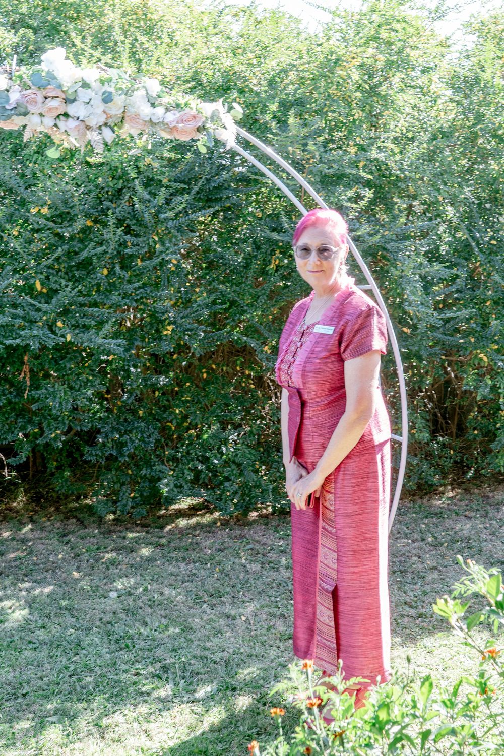 wedding celebrant in pink hair and suit posing beside pink and white round wedding arbour