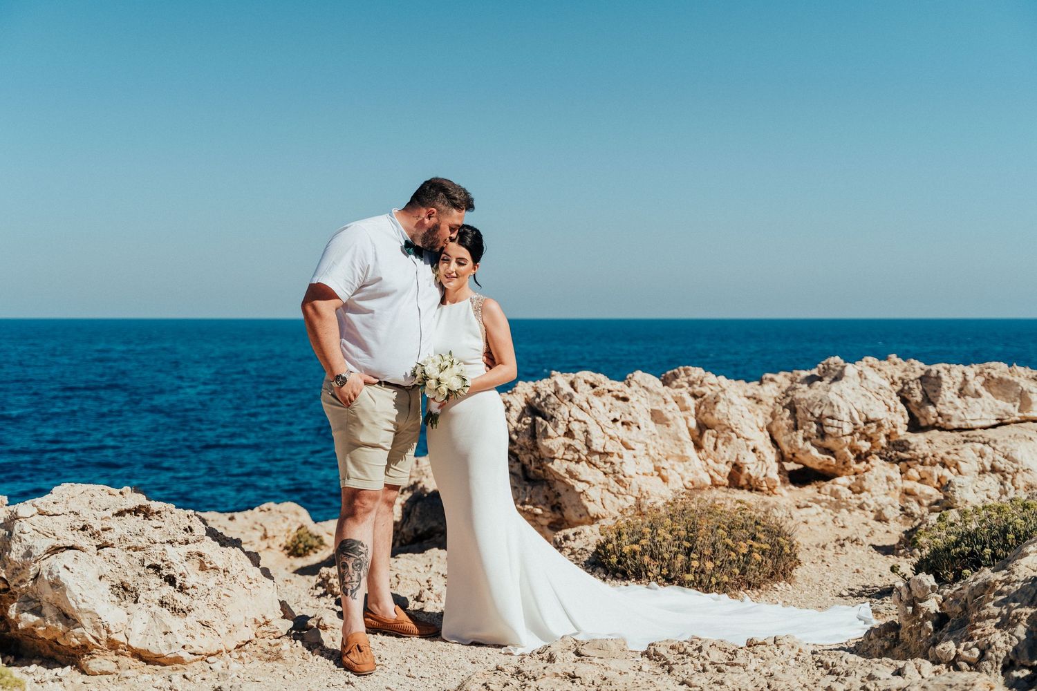 Destination wedding couple poses on rocky coastal cliff overlooking bright blue ocean.