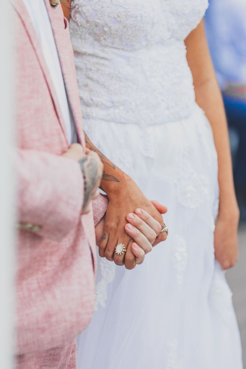 Close-up of couple holding hands showing wedding rings and attire details in pink and white.