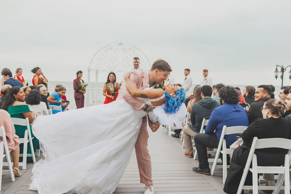 Wedding couple sharing romantic dip kiss on waterfront deck surrounded by seated guests.