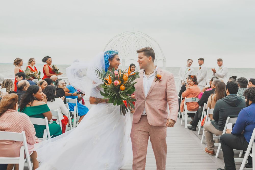 Wedding ceremony on waterfront deck with couple surrounded by seated guests and bright bouquet.