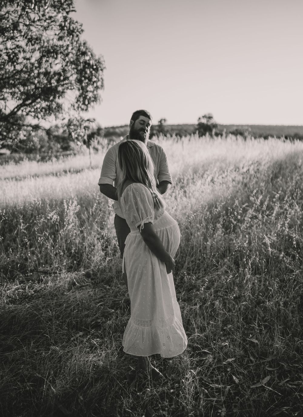 A couple in white clothing embraces in a sunlit meadow of tall grass.