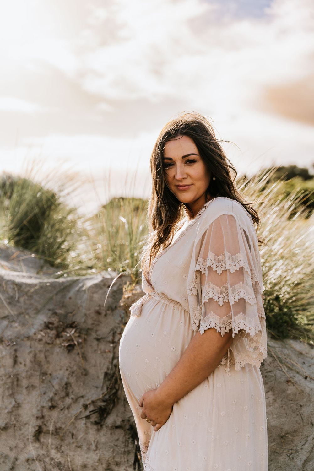 Maternity photos show woman in flowing white dress against natural beach backdrop.