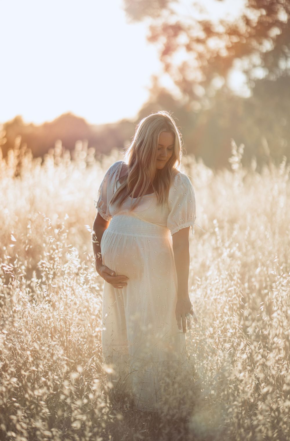 Dreamy maternity portrait in sunlit field with white wildflowers and soft bokeh effect.