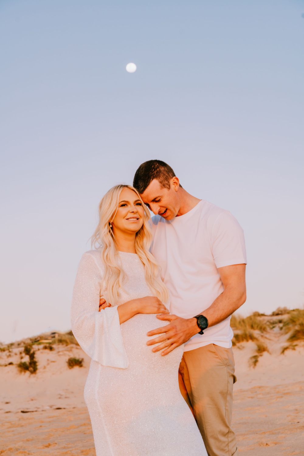 A couple shares an intimate moment on the beach with the moon visible in the sky.