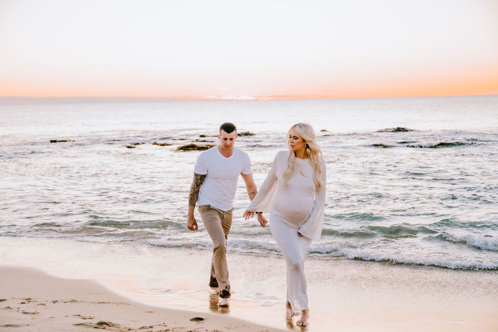 A couple walks together on a beach at sunset wearing white outfits.