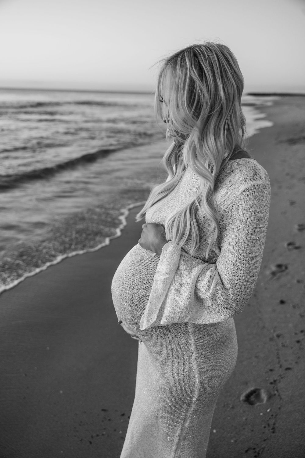 A black and white photo of a pregnant woman gazing at her baby bump on a beach.