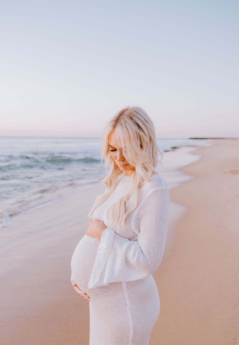 A side view of a pregnant woman in white standing on a beach at sunset.