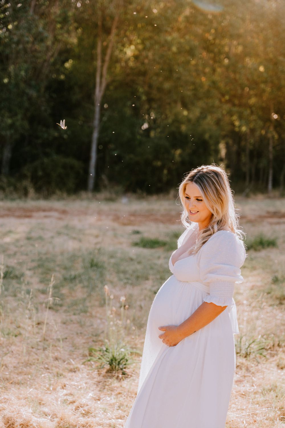 A pregnant woman in a white dress stands in a sunlit field during golden hour.