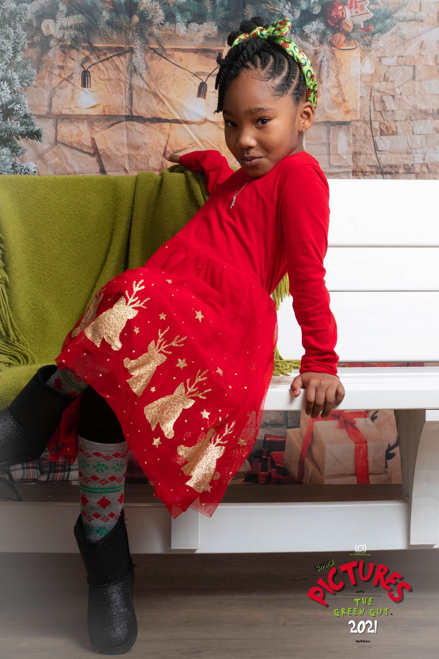 Child in festive red dress with reindeer pattern poses on white stairs next to green chair during holiday photoshoot.