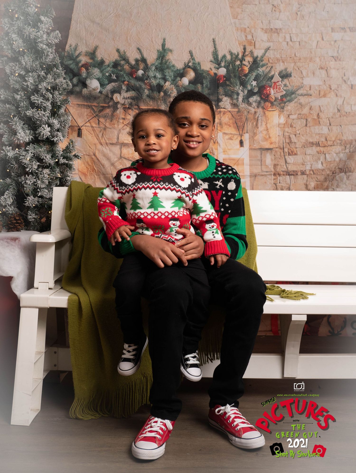 Two children wearing Christmas sweaters sit together on a white bench with holiday decorations in the background.