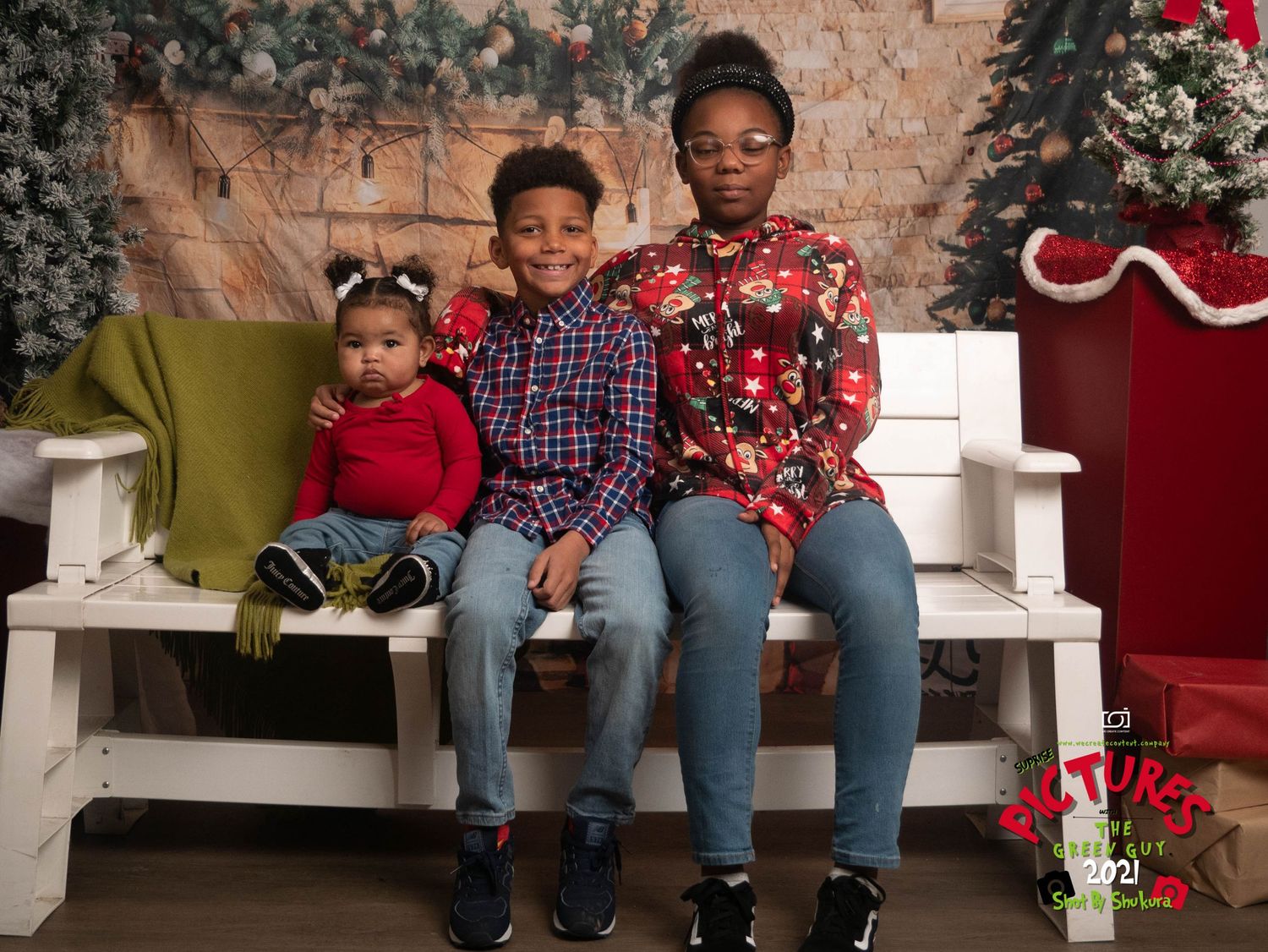 Three siblings in festive holiday outfits pose together on a white bench with winter decorations.