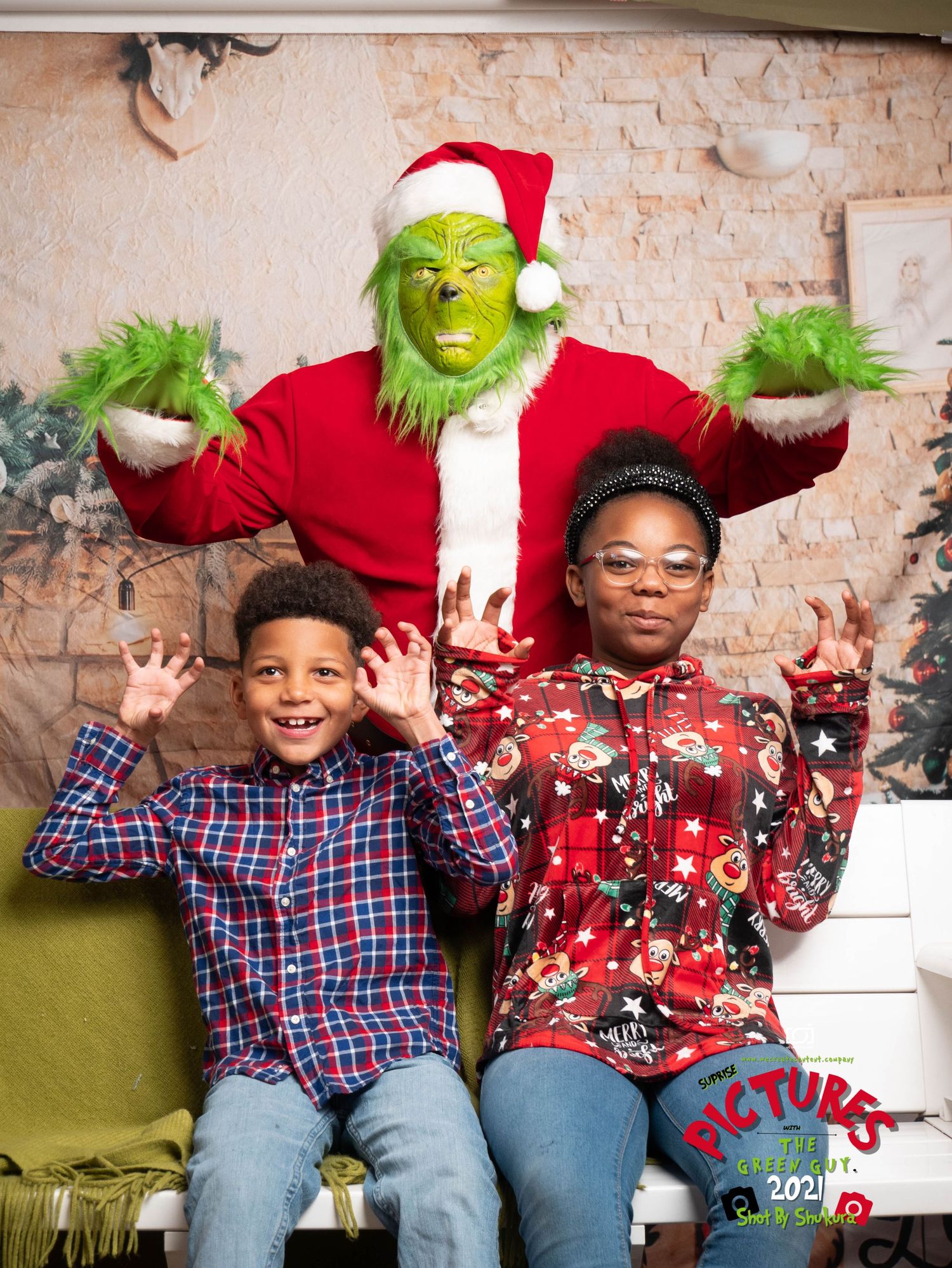 The Grinch in Santa attire poses playfully with two people against a holiday-decorated brick wall.
