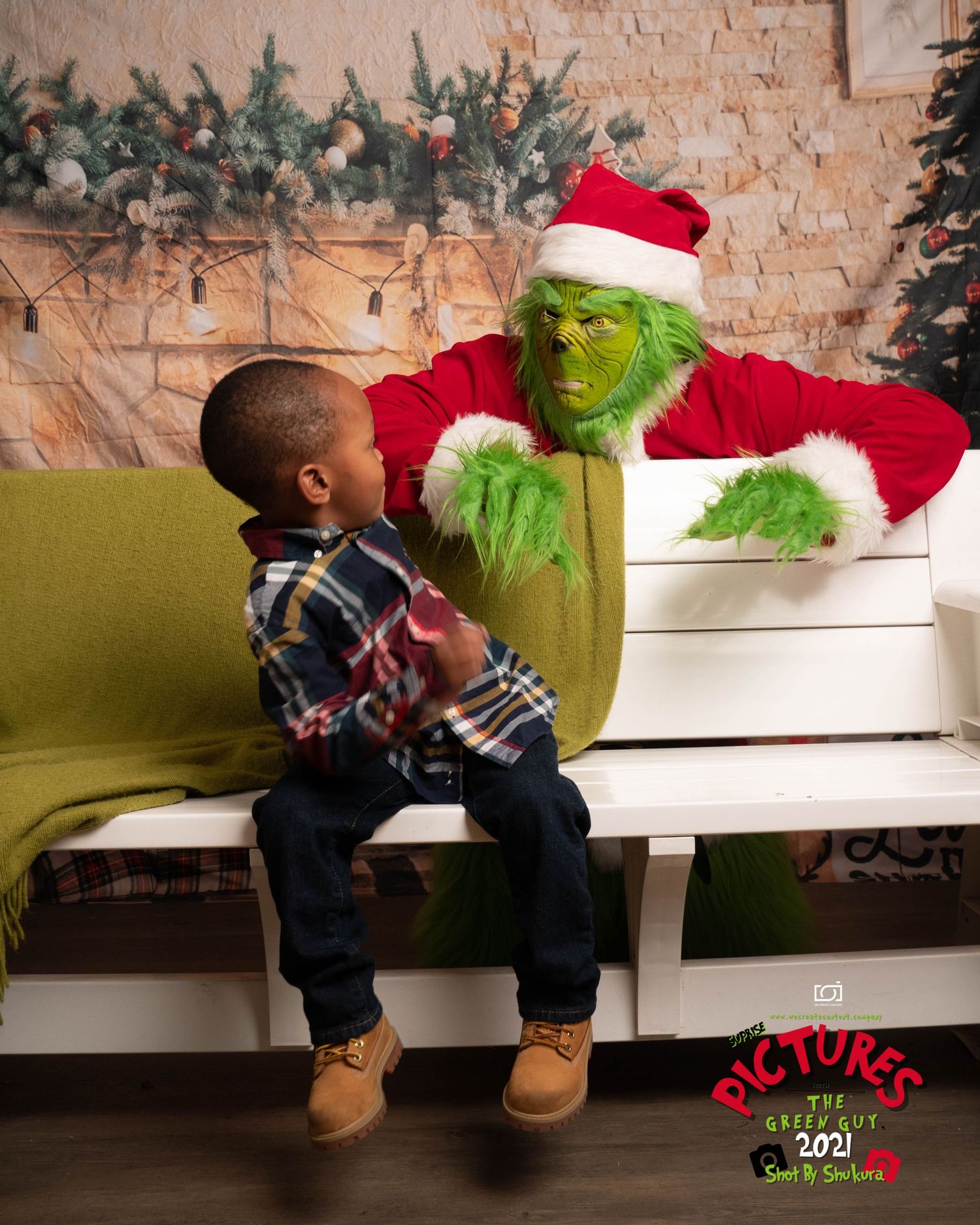 A small child sits on a festive Christmas bench looking at the Grinch character in a Santa suit against a decorated wall.