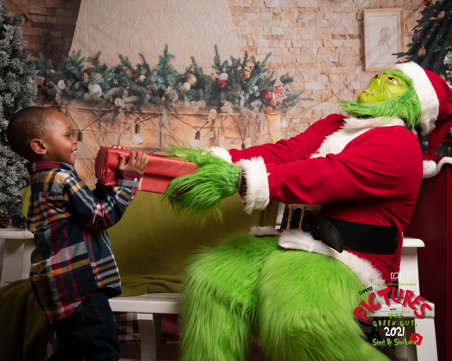 The Grinch in a Santa suit gives a present to a young child near a white brick wall with holiday decorations.