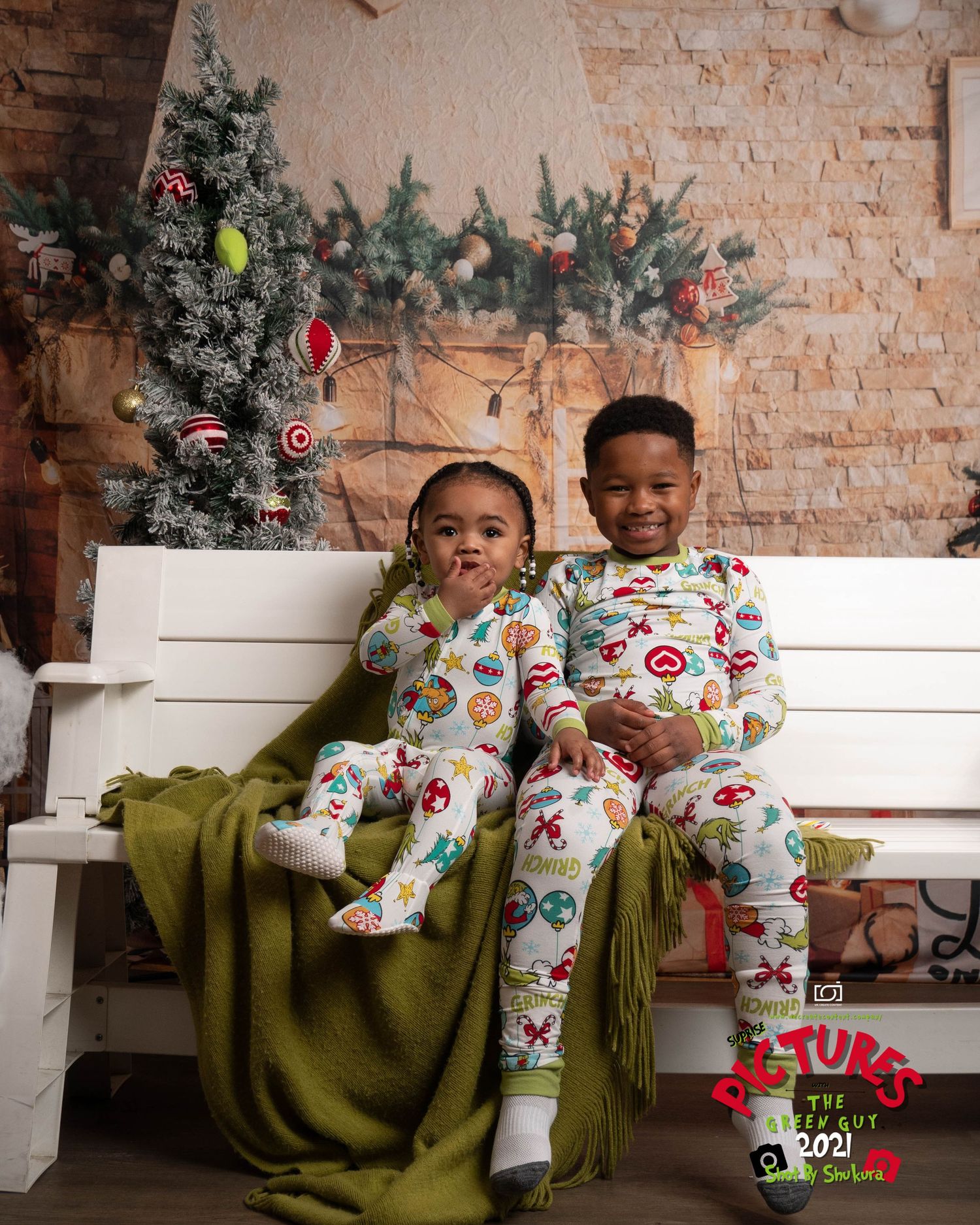 Two children in matching holiday pajamas sit on a white bench with a decorated Christmas tree in the background.