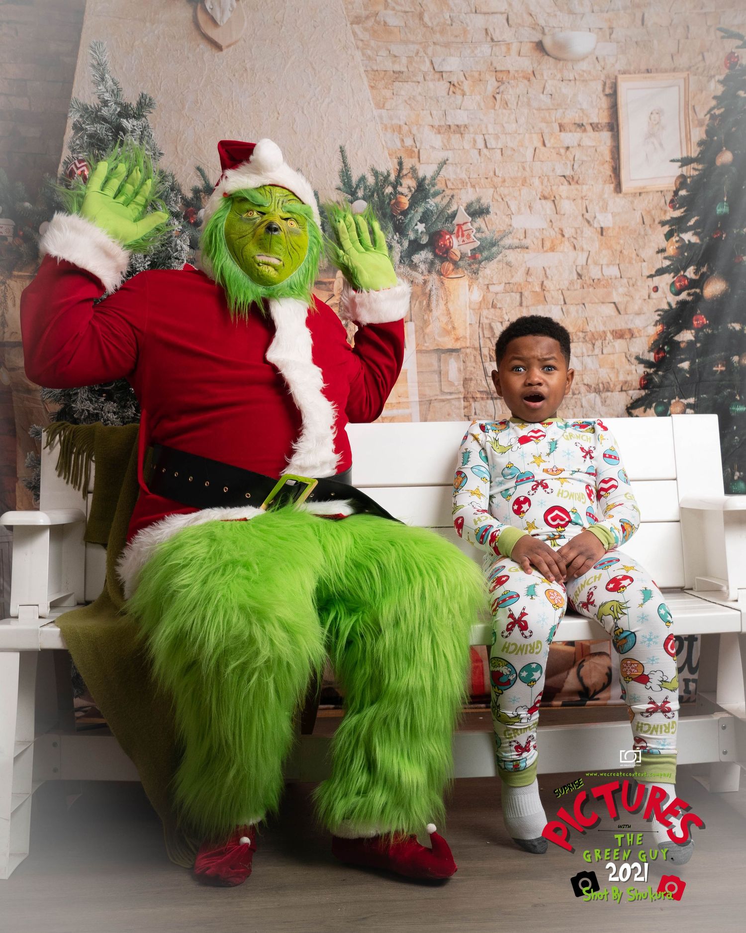 A person wearing holiday pajamas sits next to a green furry character in a Santa suit on a white bench against a brick wall.
