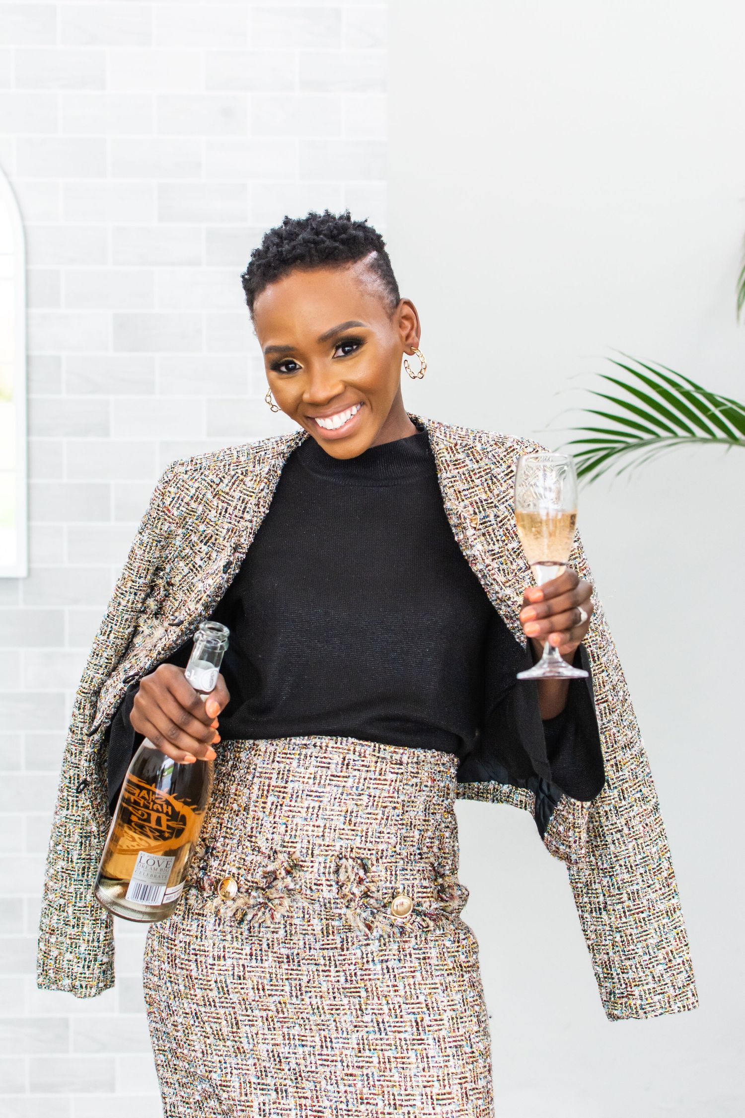 Professional woman in stylish tweed suit holding champagne and liquor bottle against white wall with palm leaf accent.