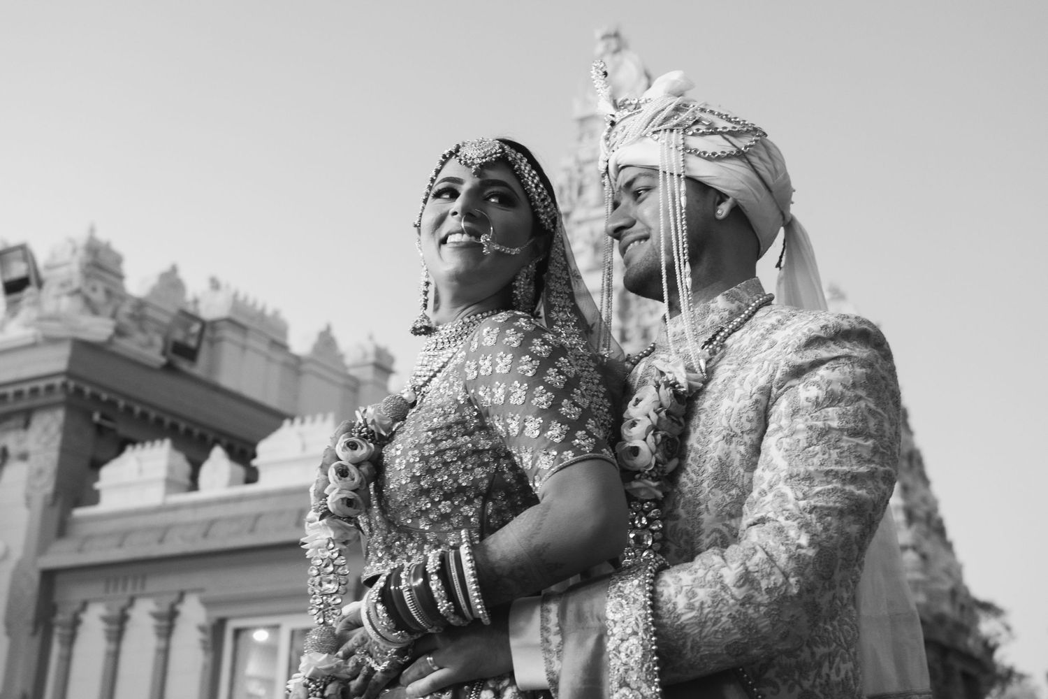 Traditional Indian wedding couple in ornate outfits pose for black and white portrait.