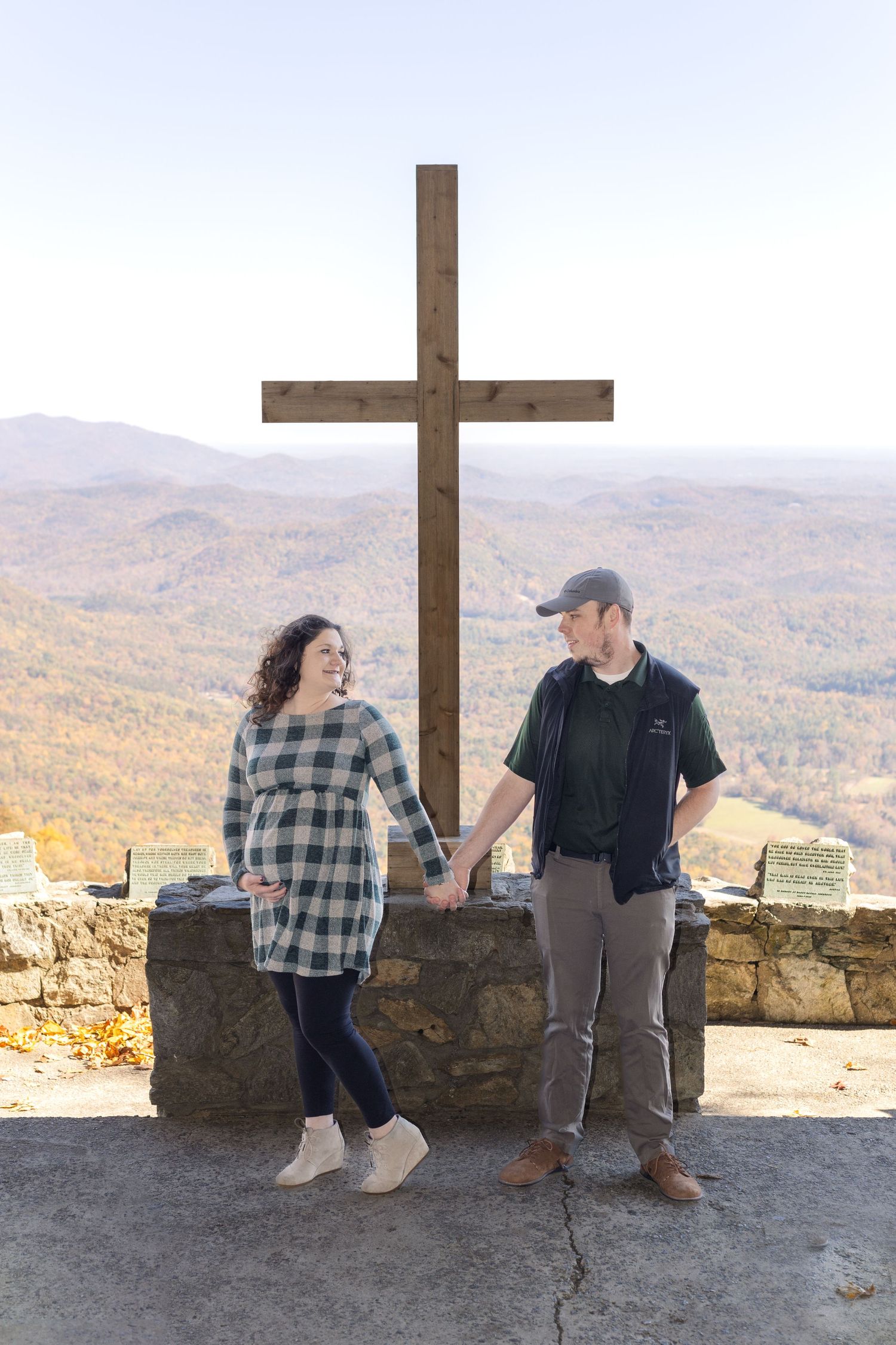 A couple poses by a large cross at a scenic mountain overlook during fall.