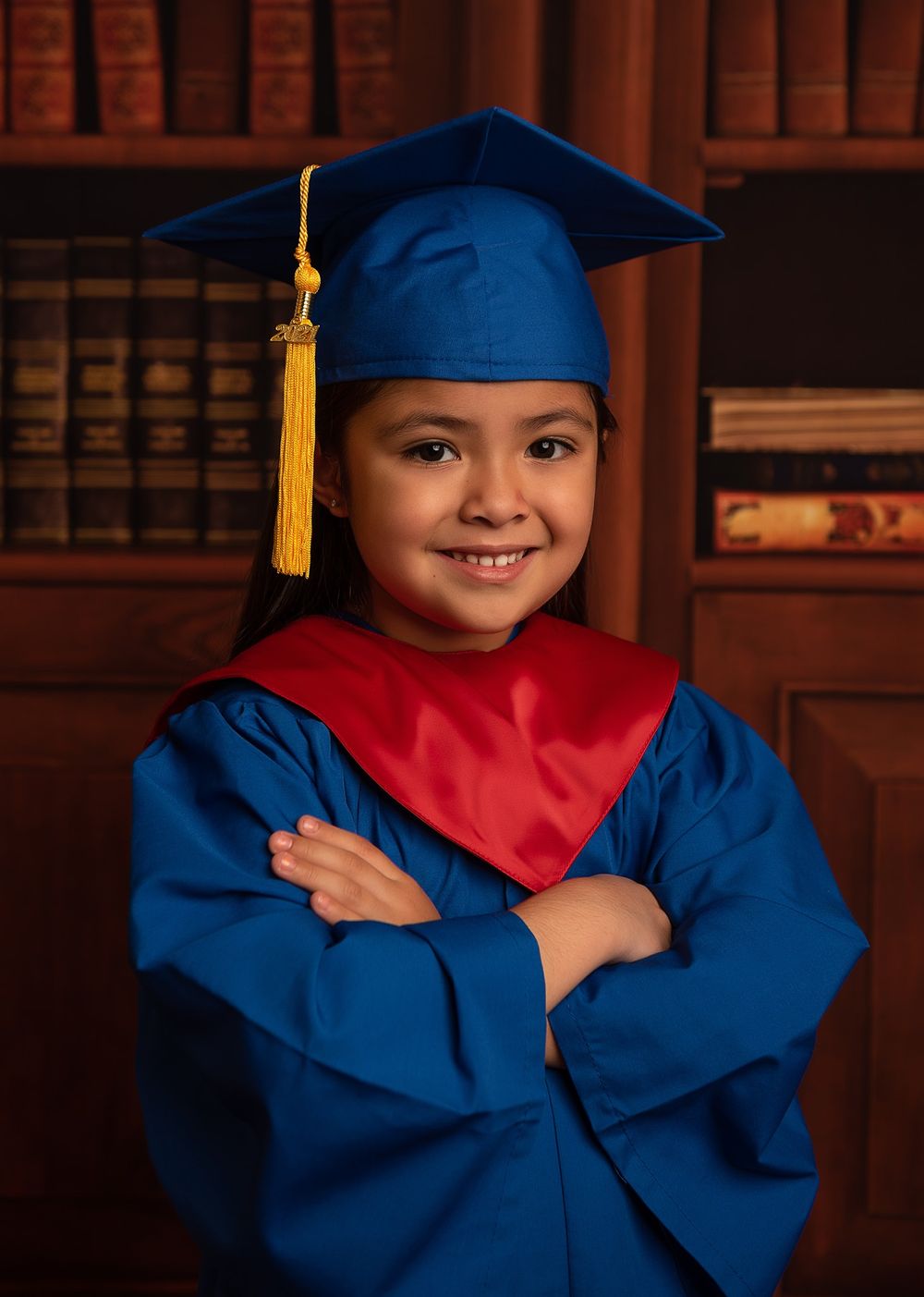 Student in blue graduation cap and gown poses confidently against library backdrop.