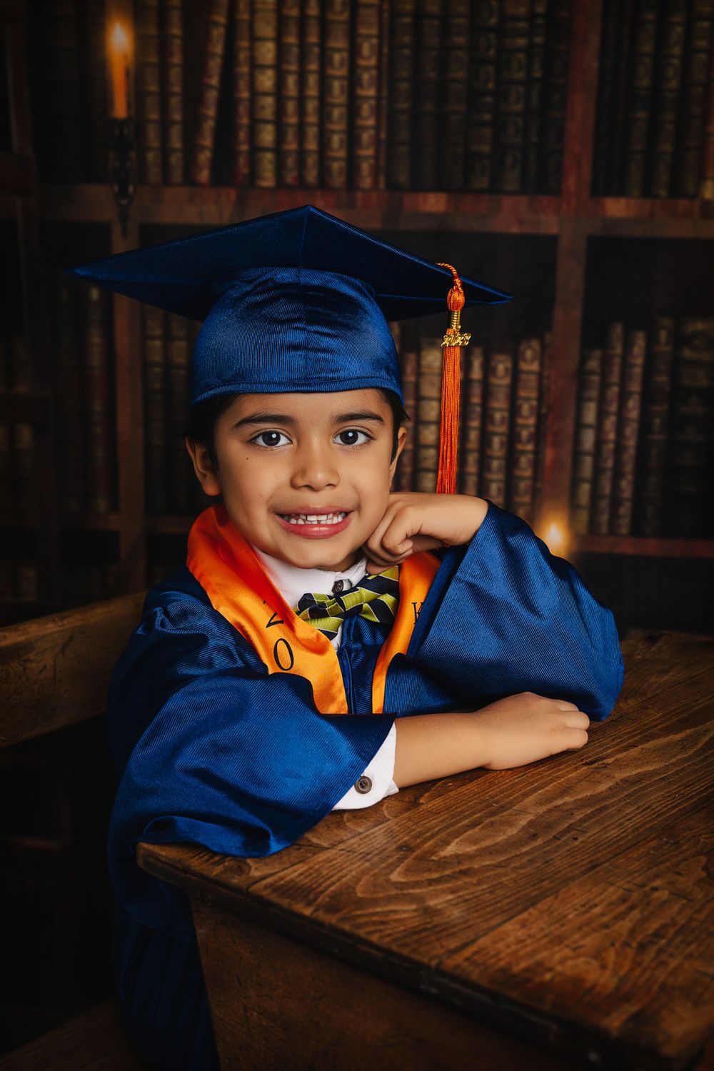 Graduate wearing blue cap and gown with orange stole poses at wooden desk in library.