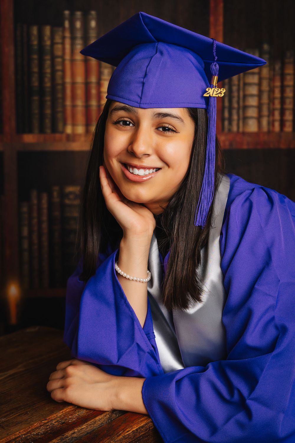 Graduate in purple cap and gown poses for portrait in front of library bookshelves.