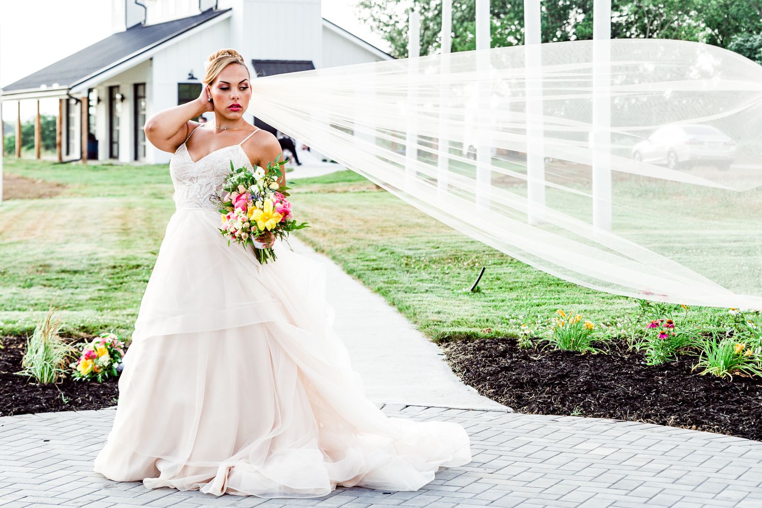 Bride in flowing white gown with long veil holds bouquet outside white fence venue by Stephanie Coulter Photography
