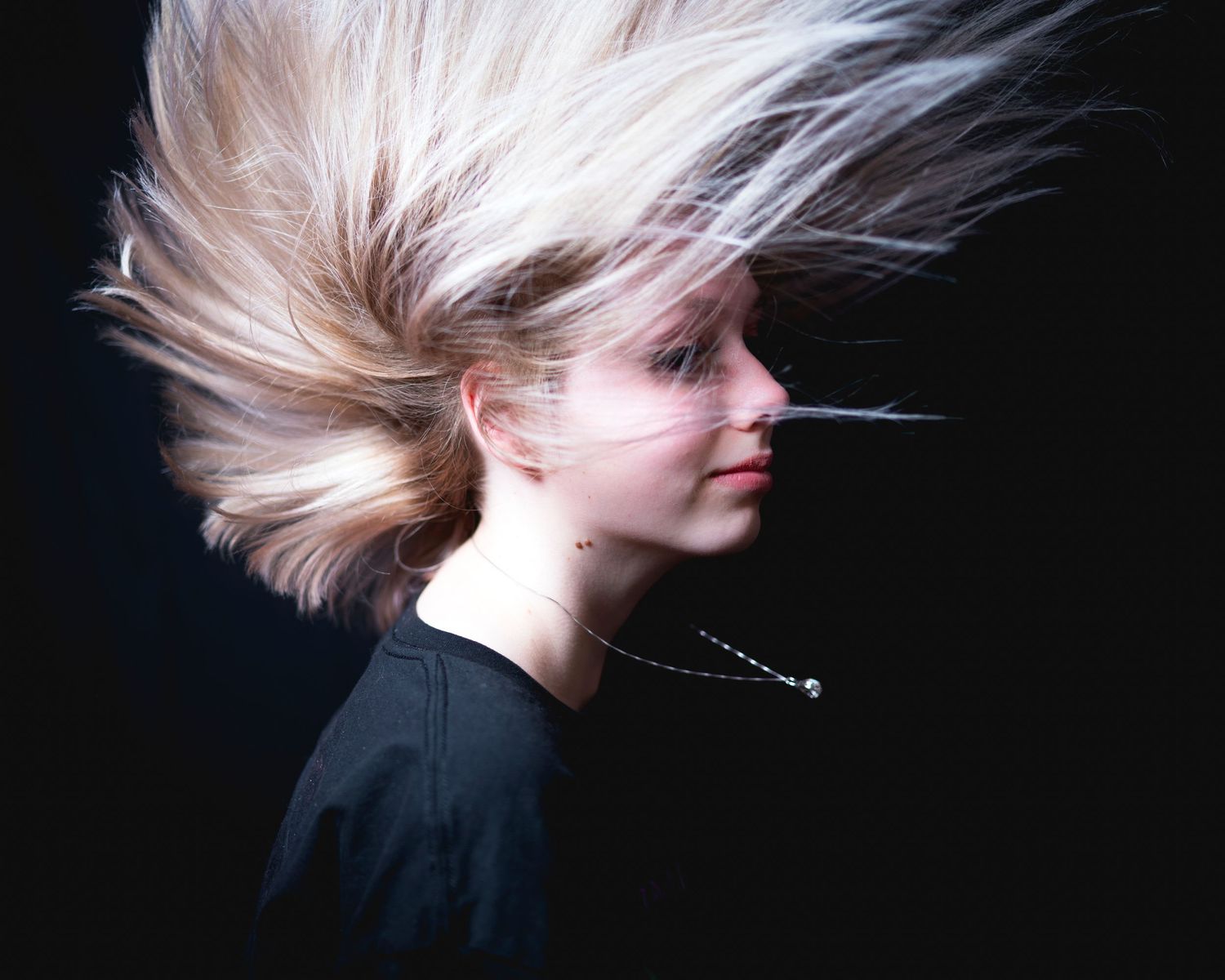 Blonde woman with hair dramatically blowing upward against dark background in dynamic portrait.