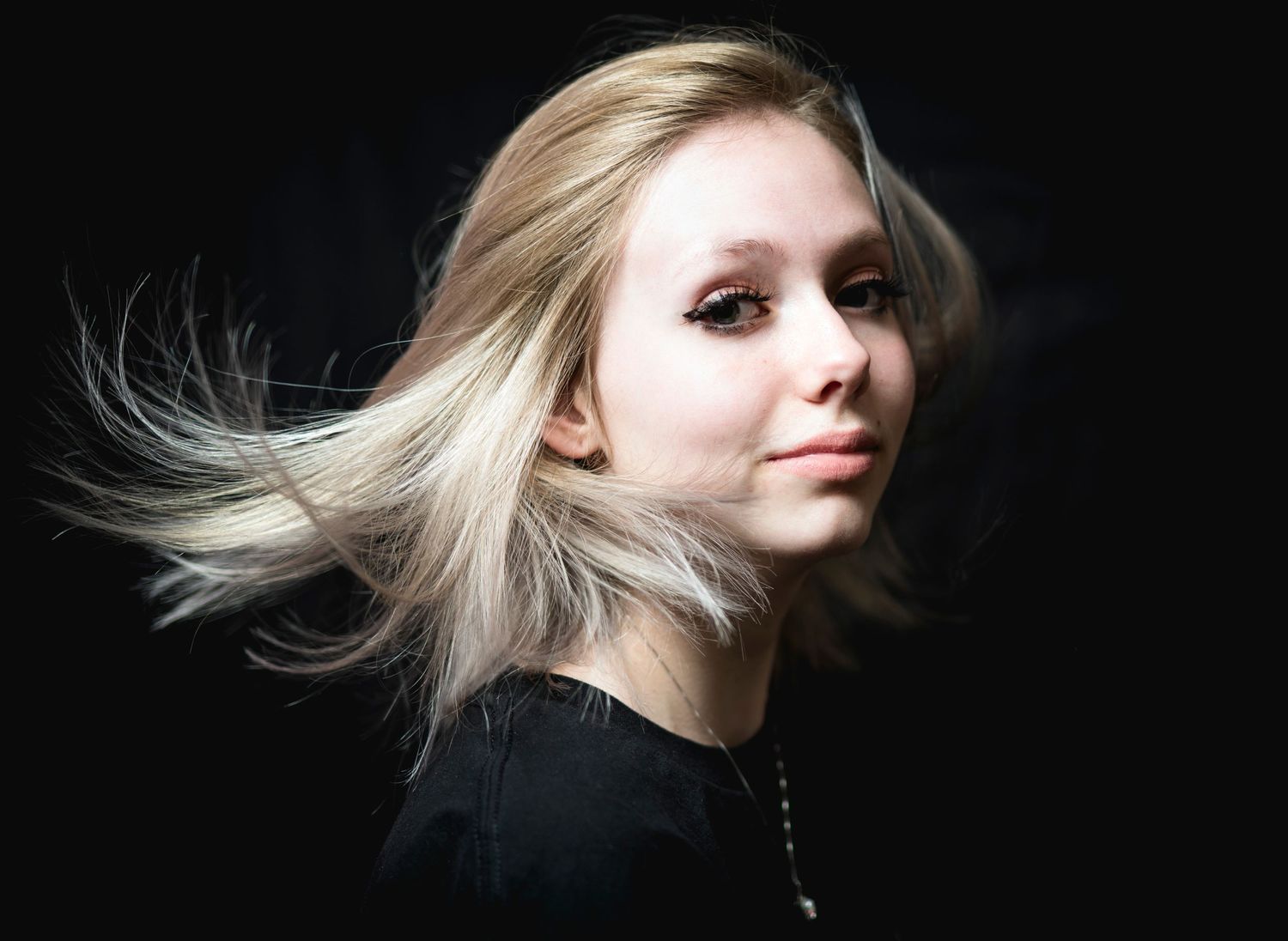 Young blonde woman with windswept hair poses against a dark background.