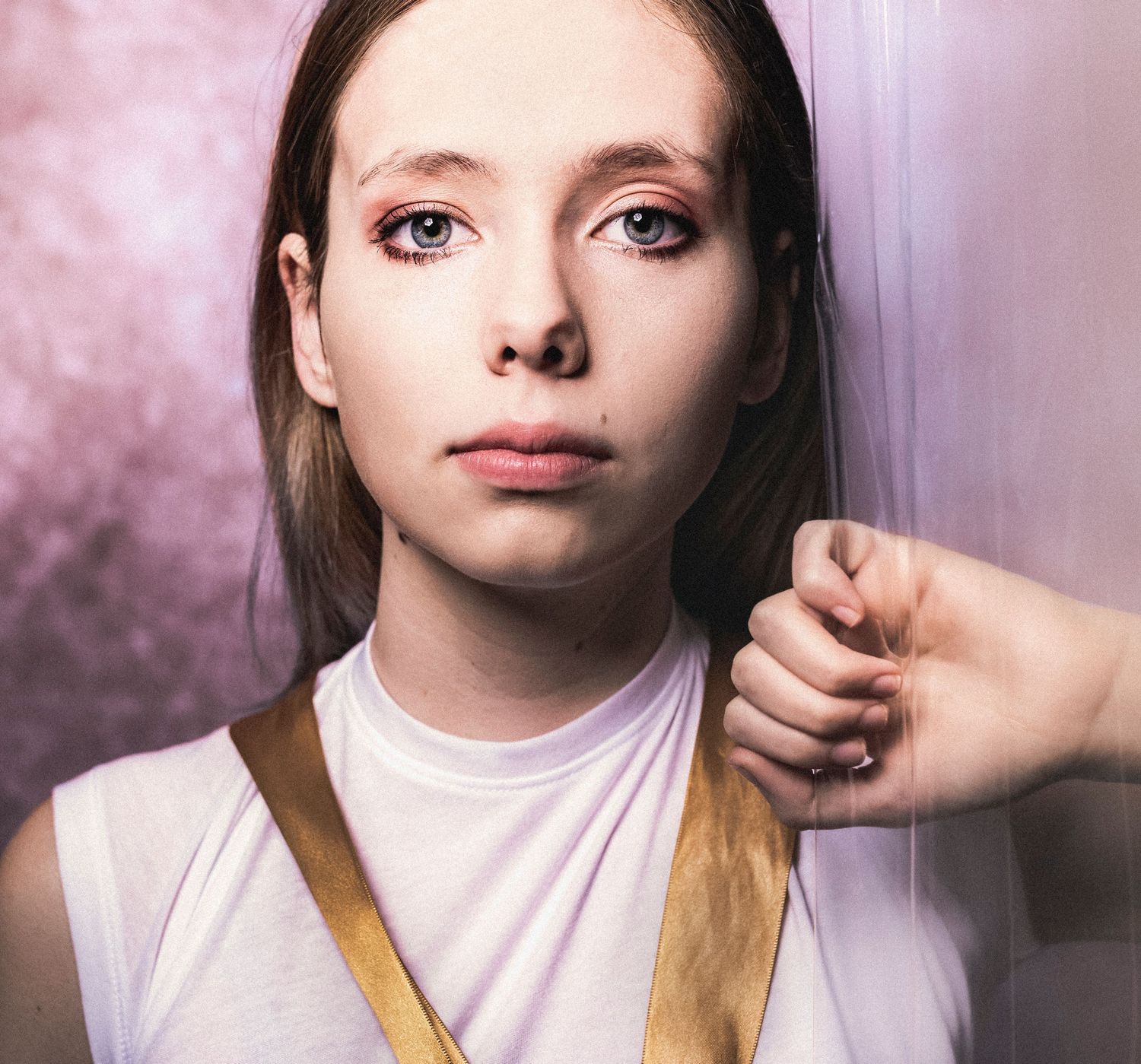 Young woman in white top with gold strap leans against a pink textured wall.