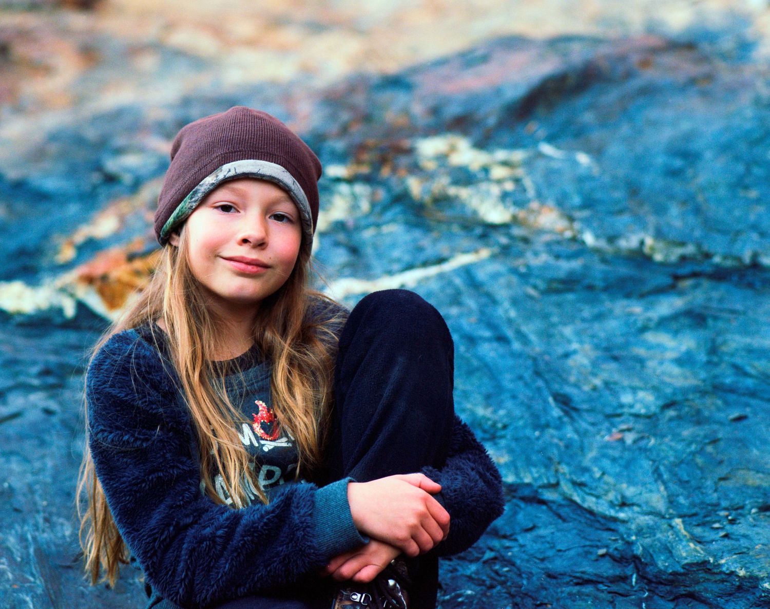 Young girl in brown beanie and blue sweater sitting on rocky surface outdoors.