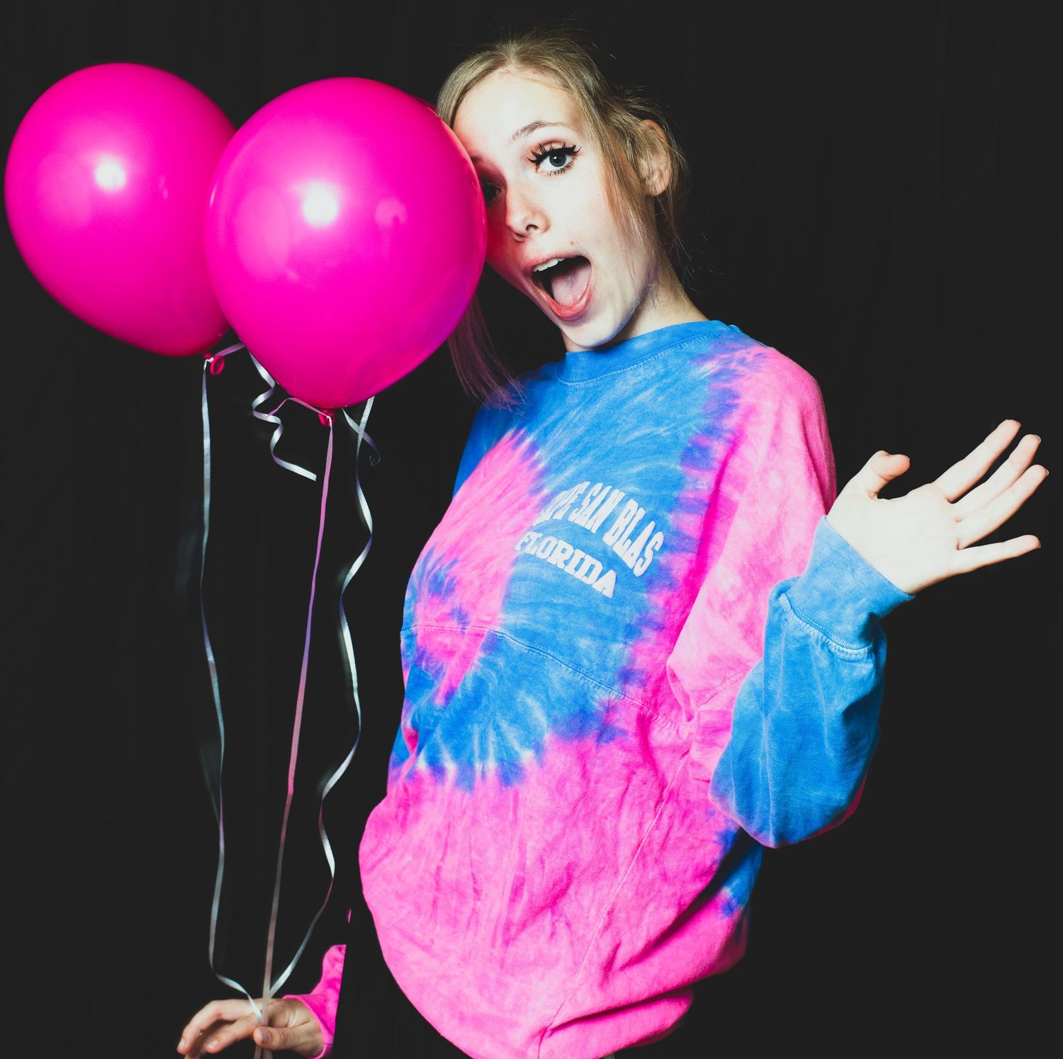 Excited blonde woman holding pink balloons wearing colorful tie-dye sweatshirt on dark background.