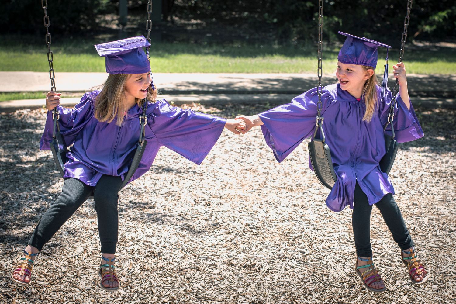 Two girls in purple graduation caps and gowns hold hands on playground swings.