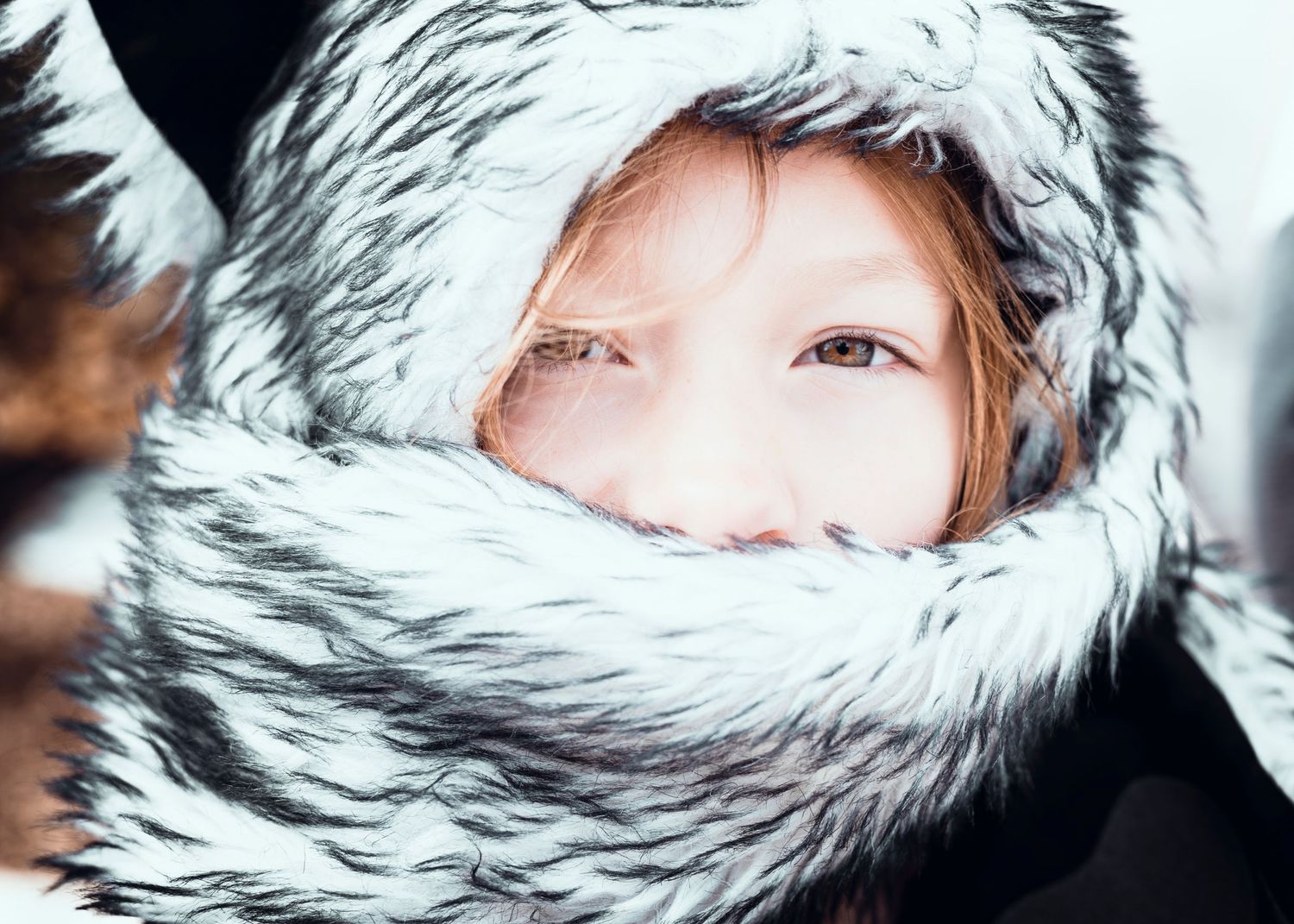 Child bundled in fur-trimmed winter hood with only eyes visible in snowy setting.