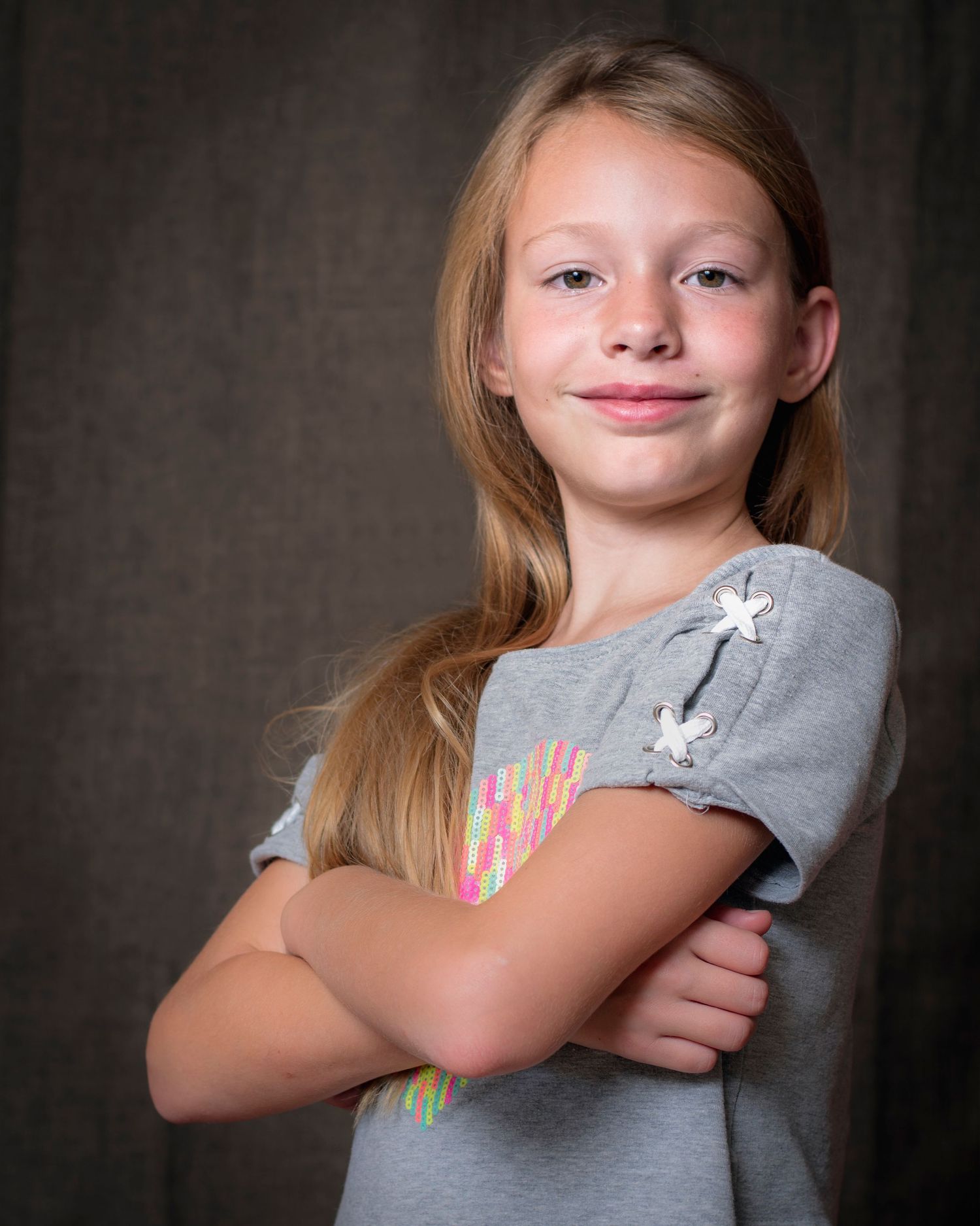 Young blonde girl in gray t-shirt posing with arms crossed against studio backdrop.