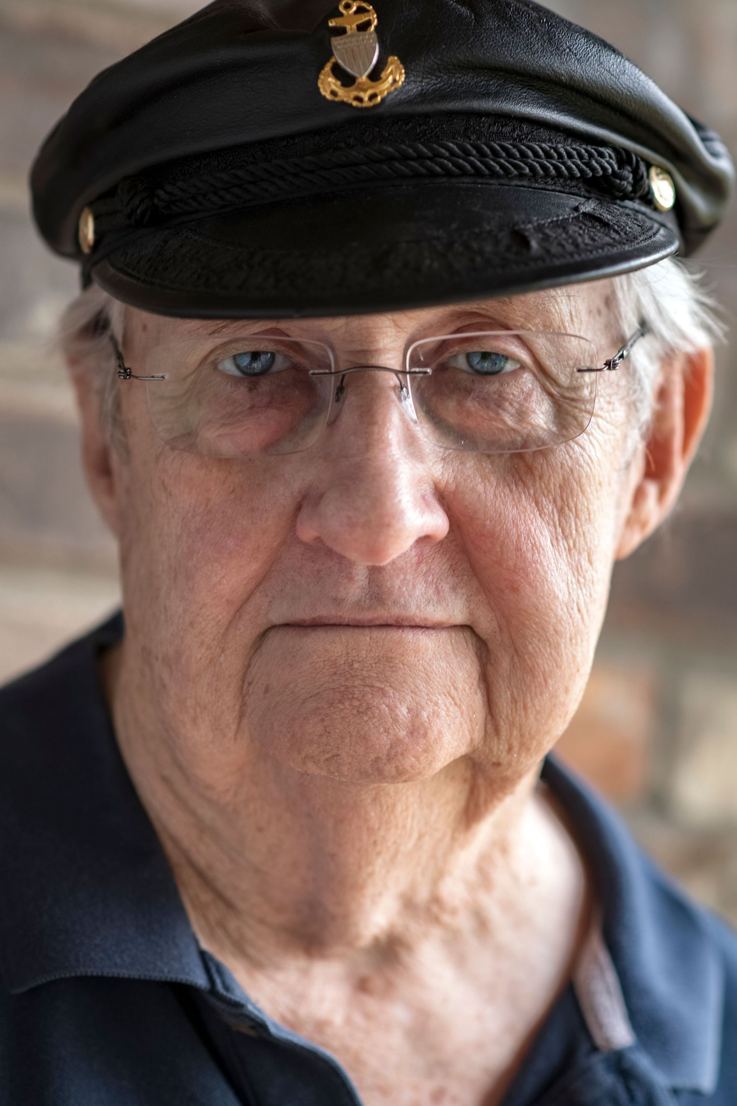Serious elderly man wearing black captain hat and glasses against brick wall background.
