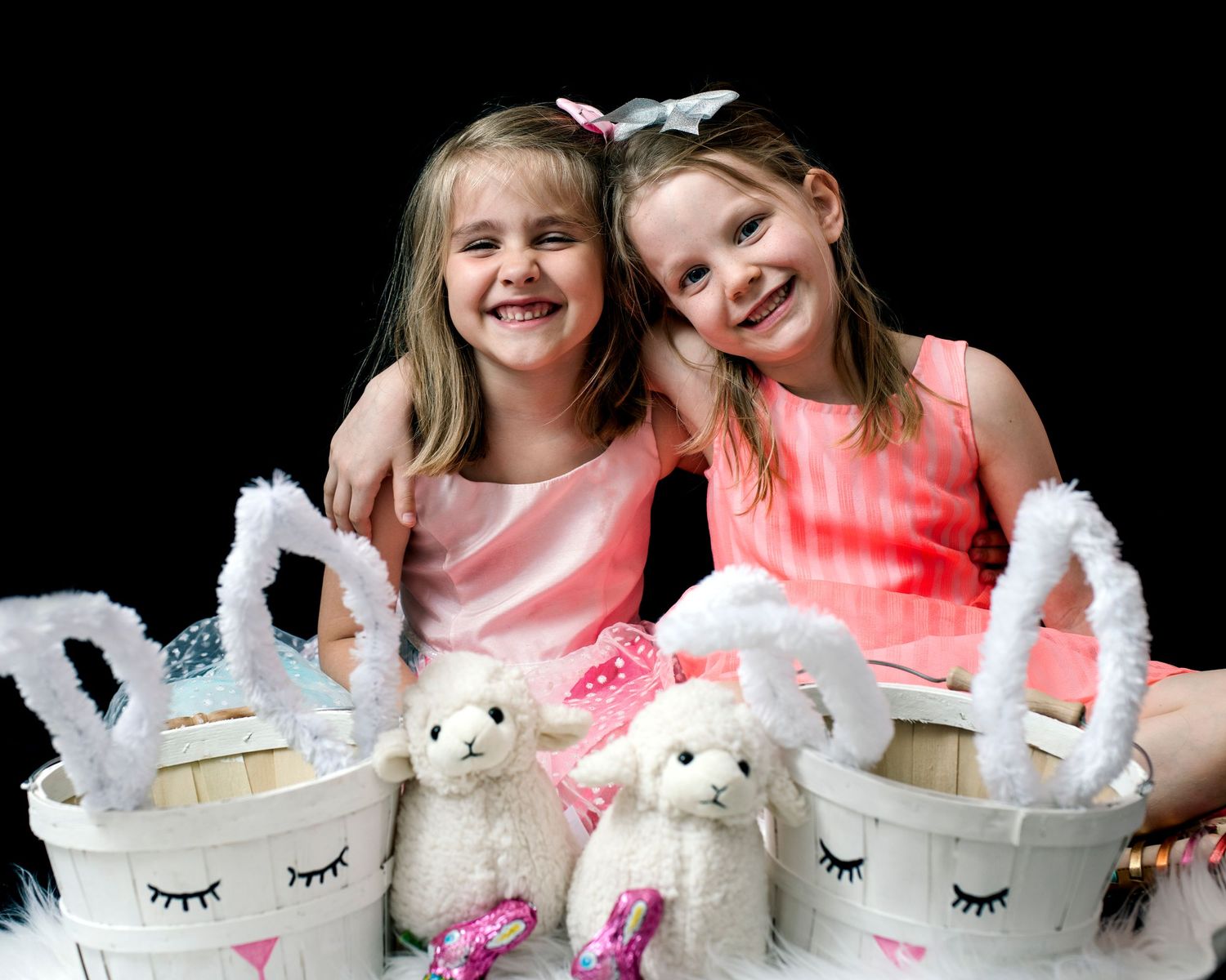 Two smiling girls in pink dresses pose with Easter baskets and stuffed lamb toys.