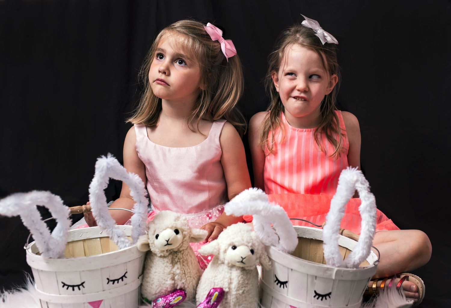 Two young girls in pink dresses sitting with Easter baskets and stuffed animal toys.