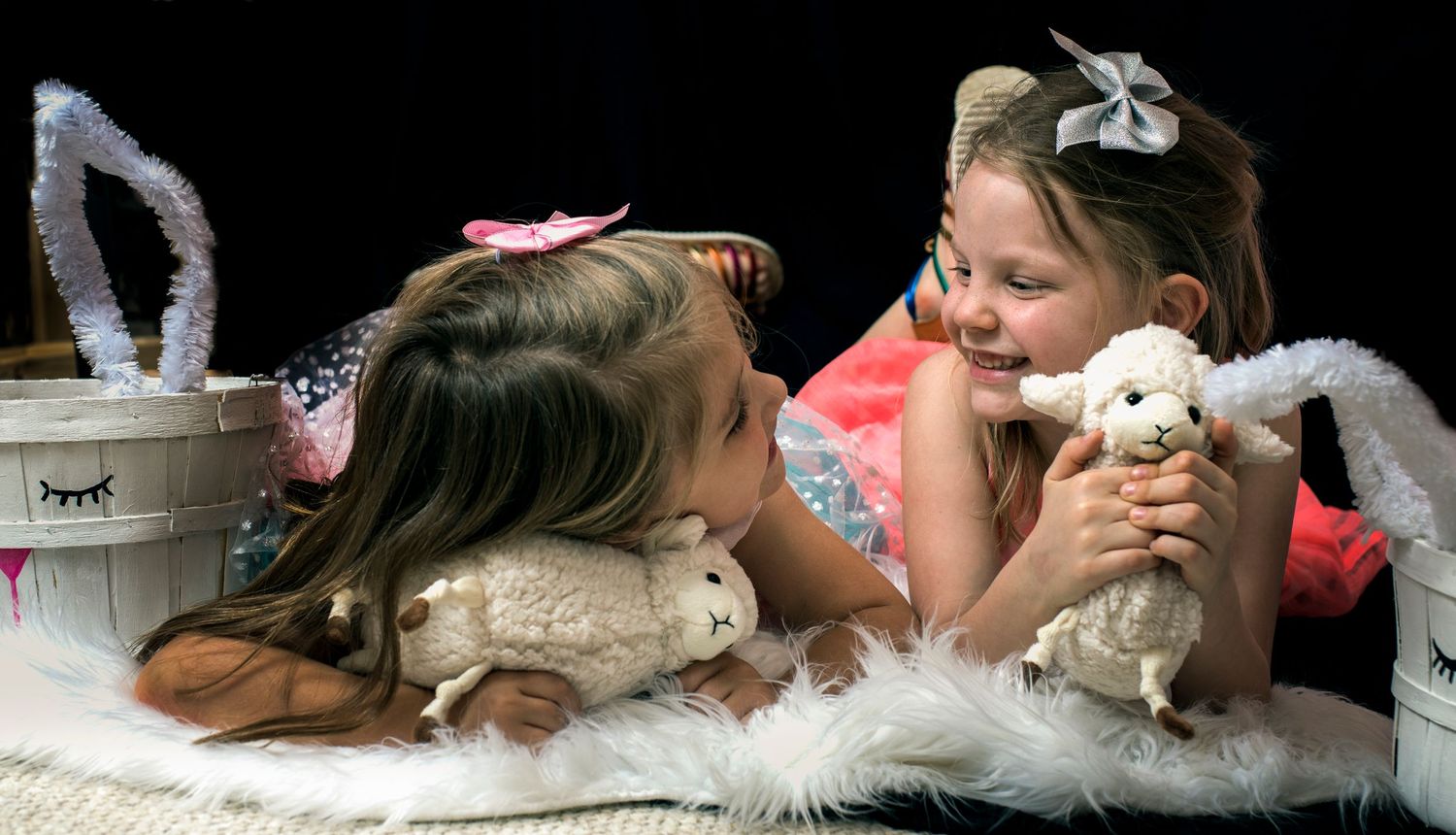 Two young girls in pink dresses lying down smiling and holding stuffed lamb toys.
