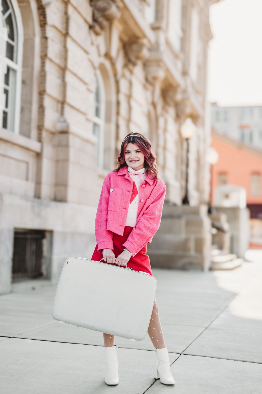 Urban fashion portrait with bright pink jacket and white boots against stone architecture.