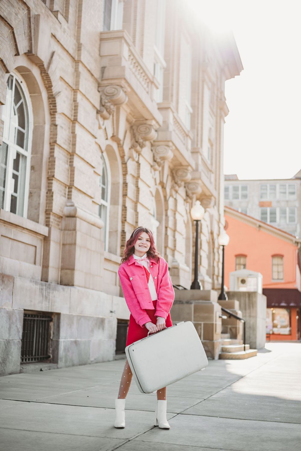 Street fashion photography with pink jacket and white handbag against historic building facade.