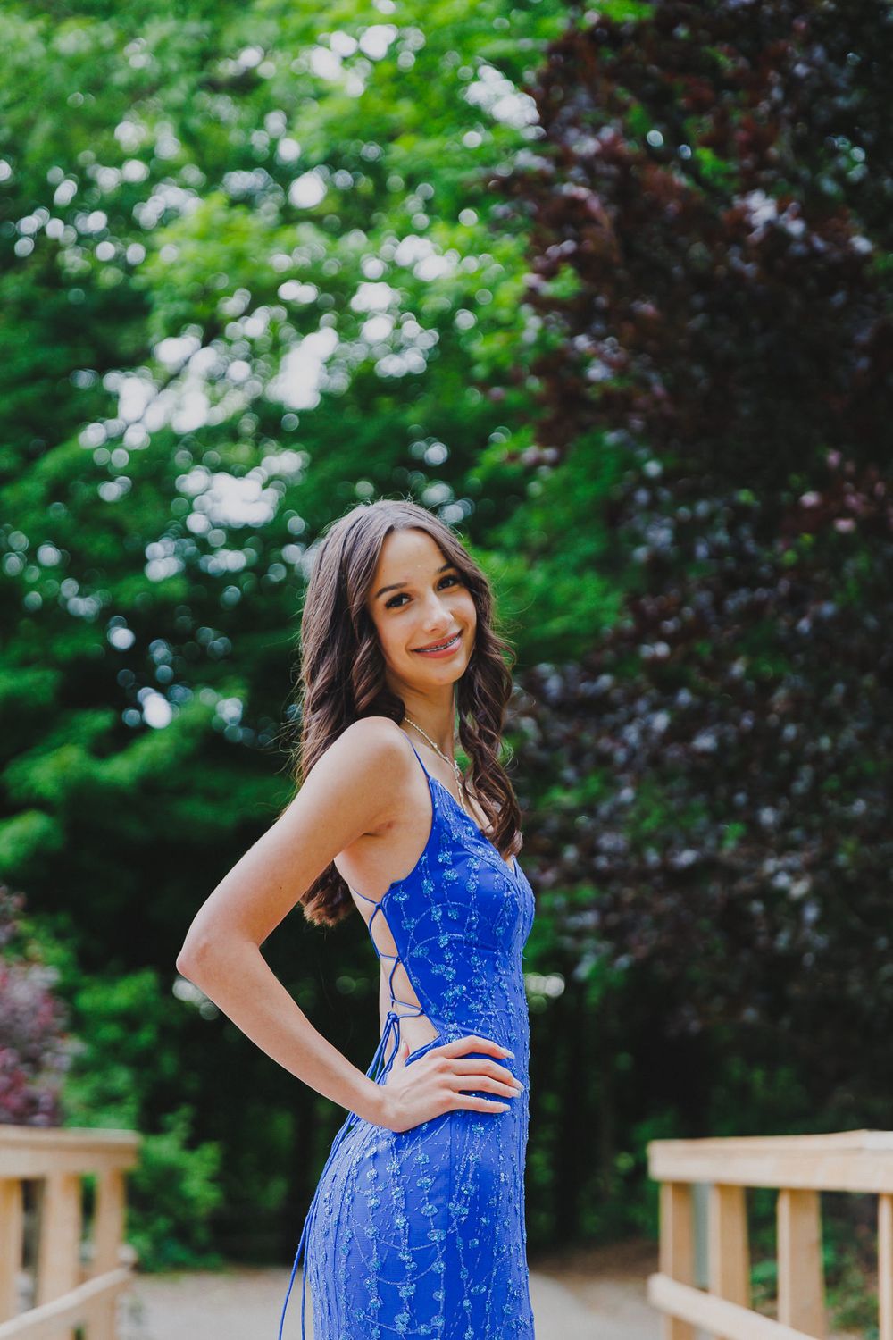 Elegant blue lace gown worn on wooden bridge deck surrounded by lush green foliage in outdoor setting.